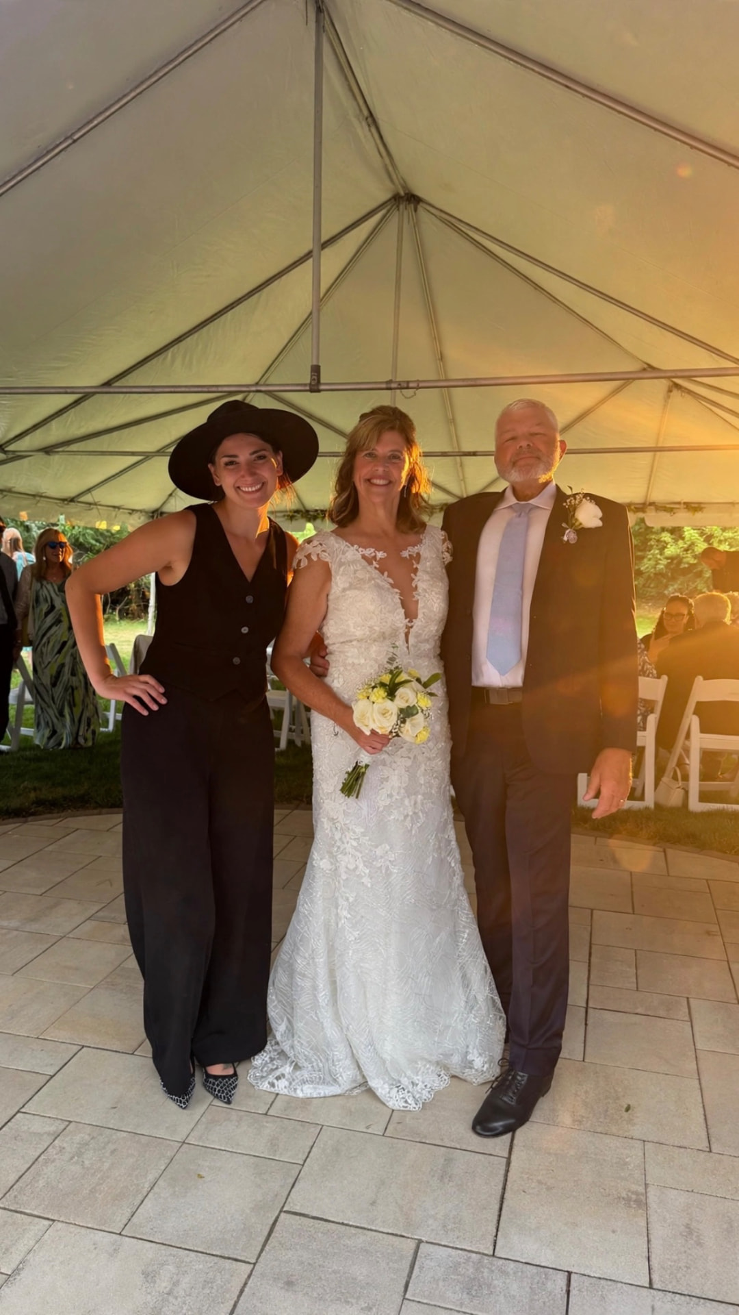 Anna Marie in a black hat with a bride and groom under a tent at sunset