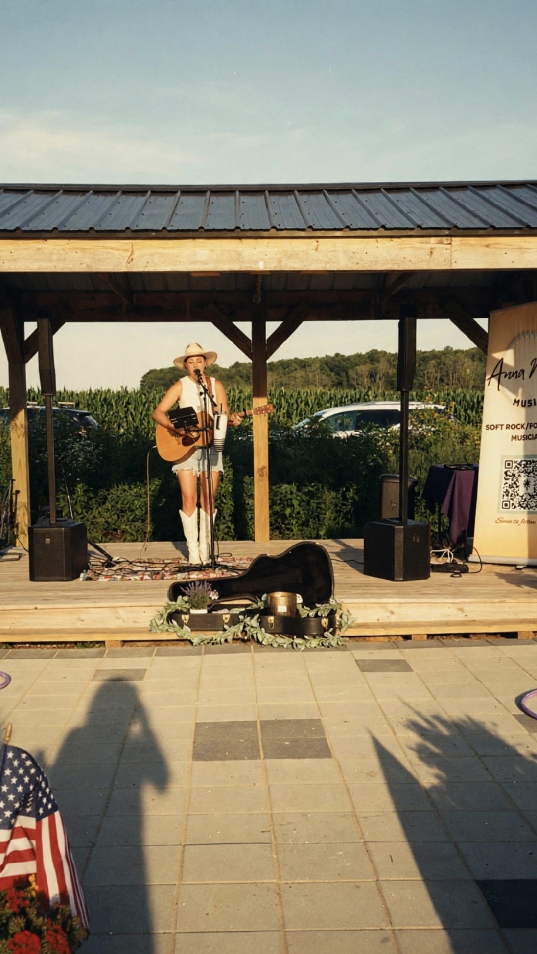 Anna Marie performing with guitar on an open-air stage beside her banner